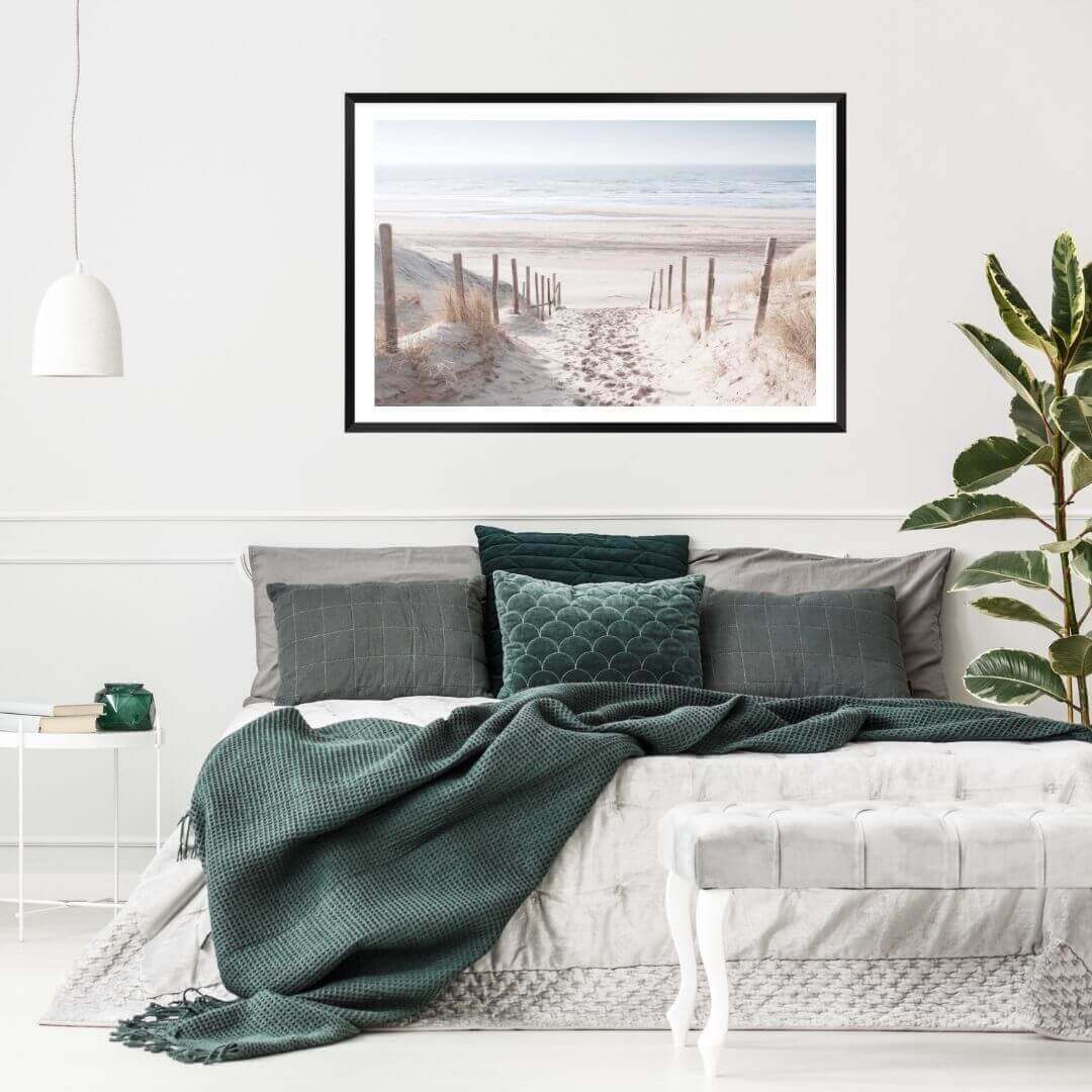 A wall art photo print of a walk on the beach with a black frame, white border on wall above bed